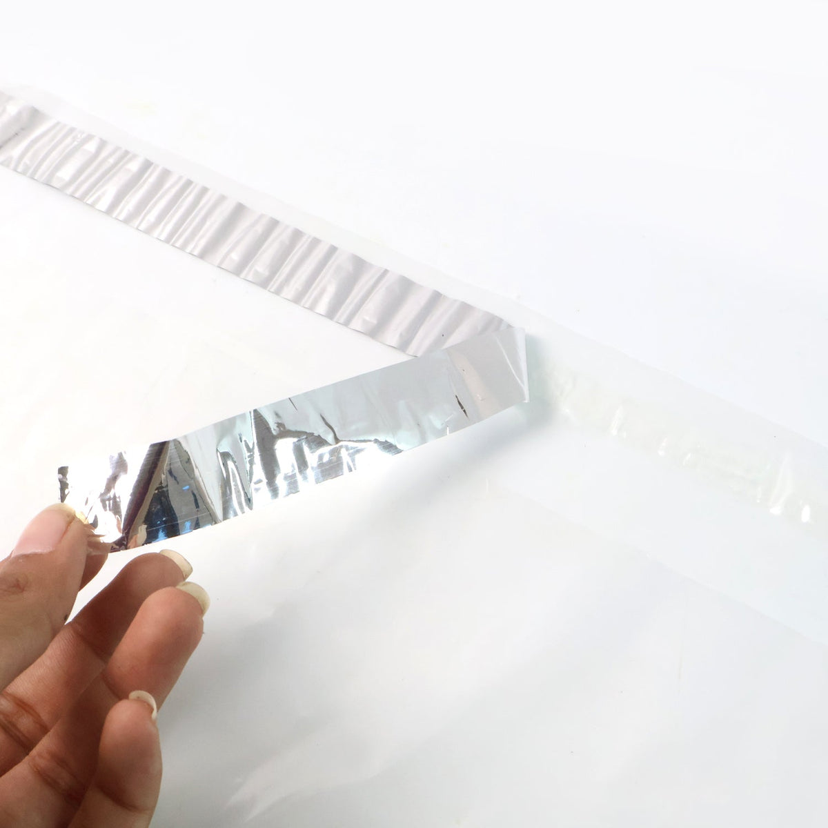 A hand peeling the silver adhesive strip off a clear polybag on a white background.