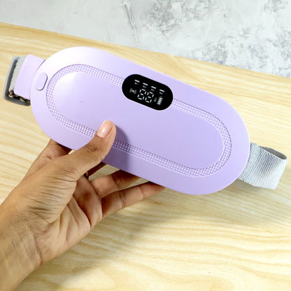 A hand holding a lavender heating pad with a digital display, against a light wooden and gray background.