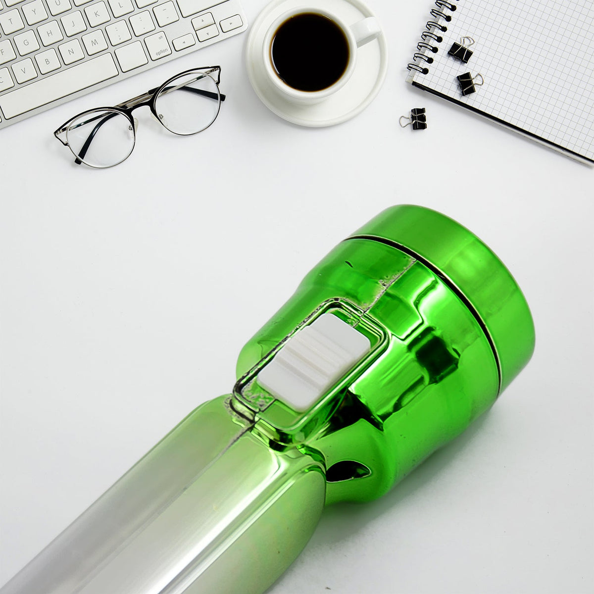 The green end of the torchlight with a white switch, on a white desk with a keyboard, coffee cup, and notebook.