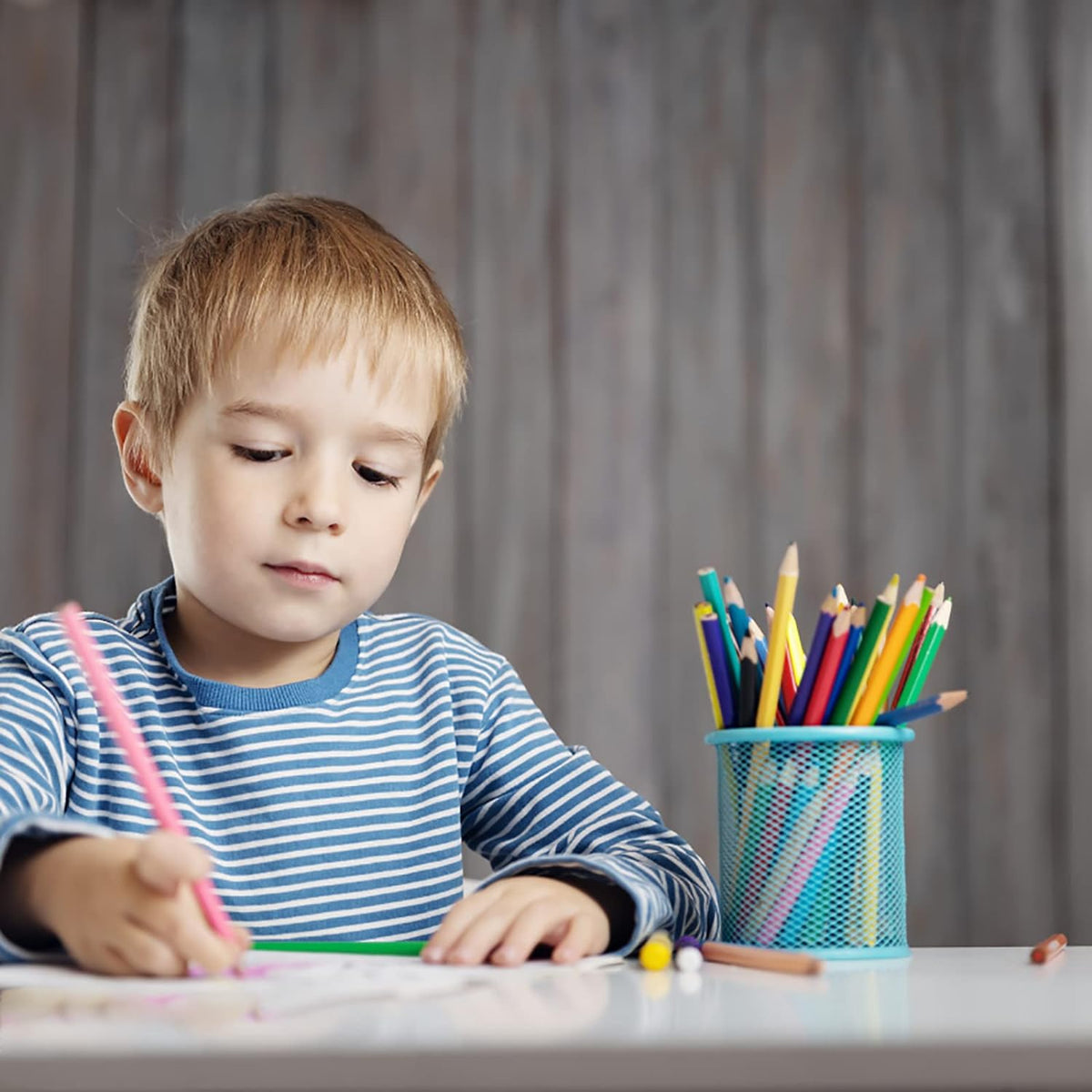 A young boy in a striped shirt draws at a white table with a blue mesh pen holder full of pencils.
