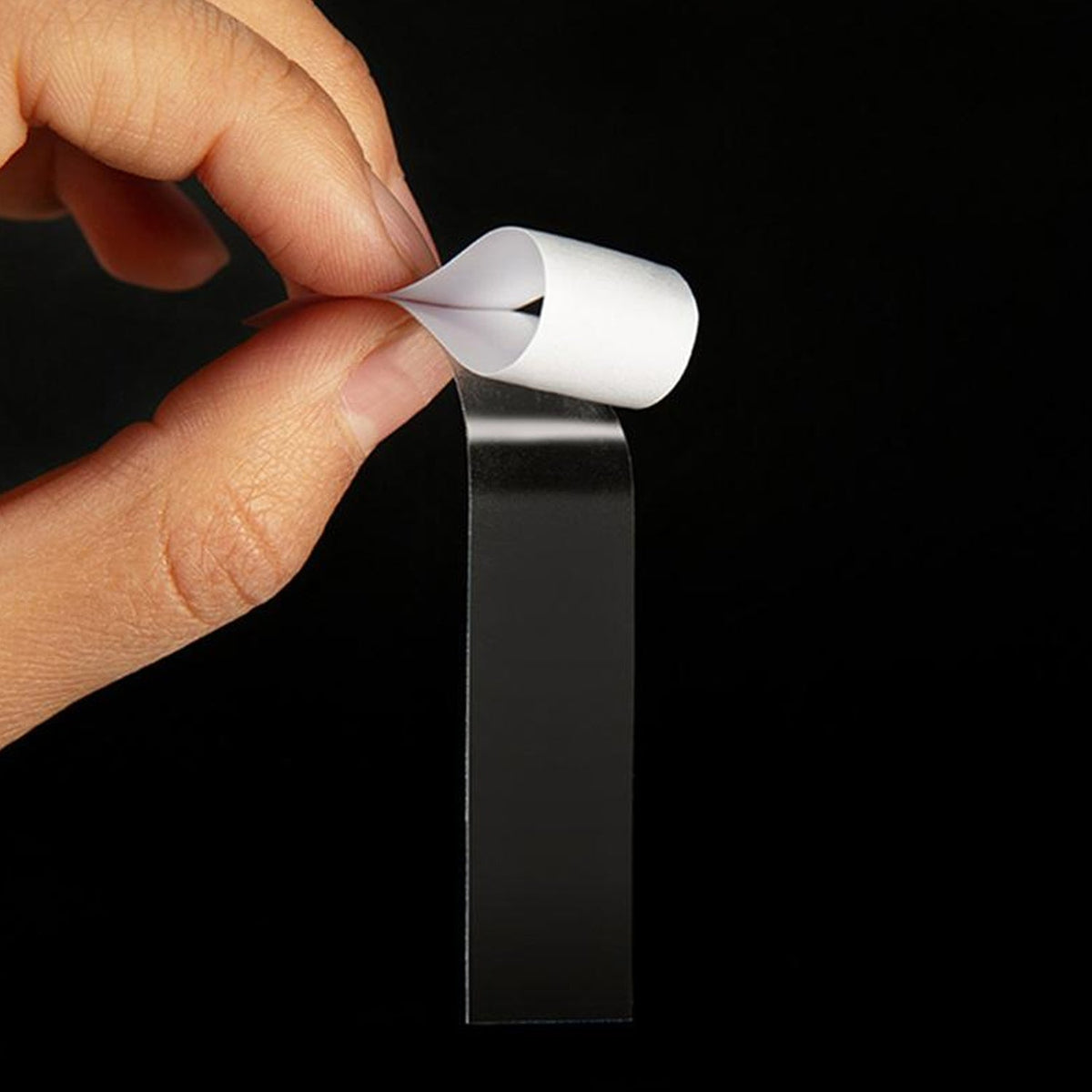 A hand holds a clear strip of tape with a white backing, peeling it away from the dark background.