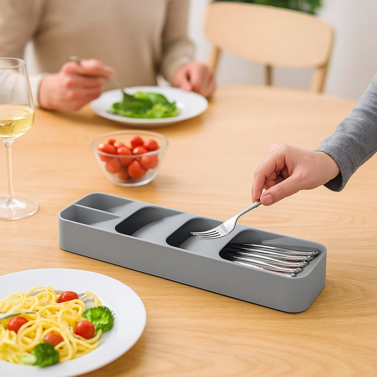A hand places a fork into a gray plastic cutlery organizer on a wooden dining table with plates of food.
