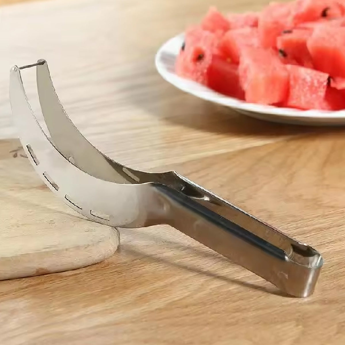 A stainless steel watermelon slicer rests on a wooden cutting board with a plate of watermelon chunks in the background.