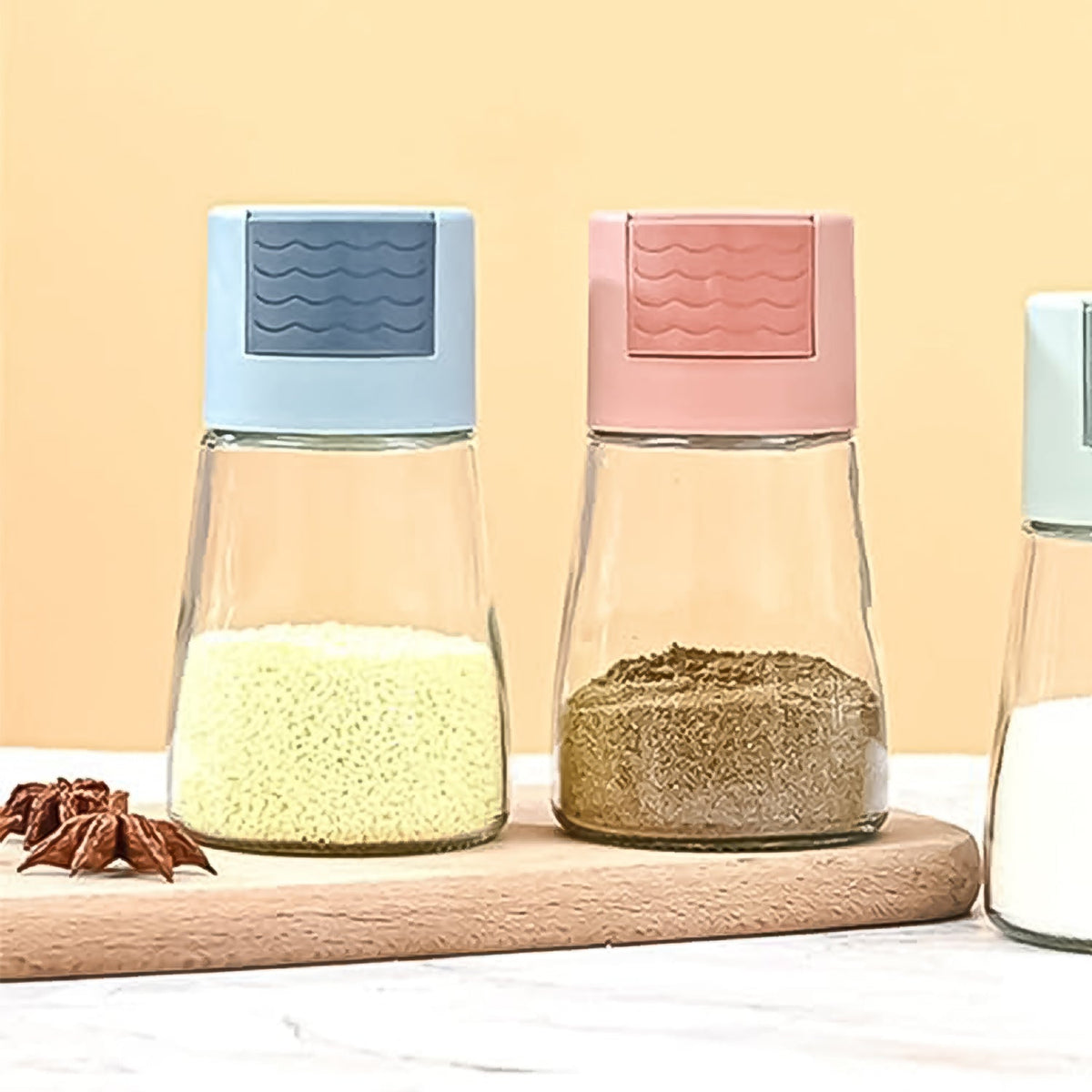 Two glass spice dispensers with colorful tops sit on a wooden board, filled with yellow and brown spices.