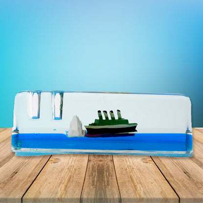 A clear rectangular liquid motion paperweight with a green ship and iceberg floats in blue liquid on a wooden surface.