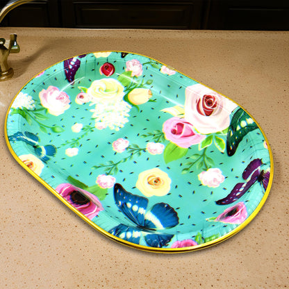 An oval turquoise tray with a floral and butterfly design rests on a light brown countertop with a dark background.