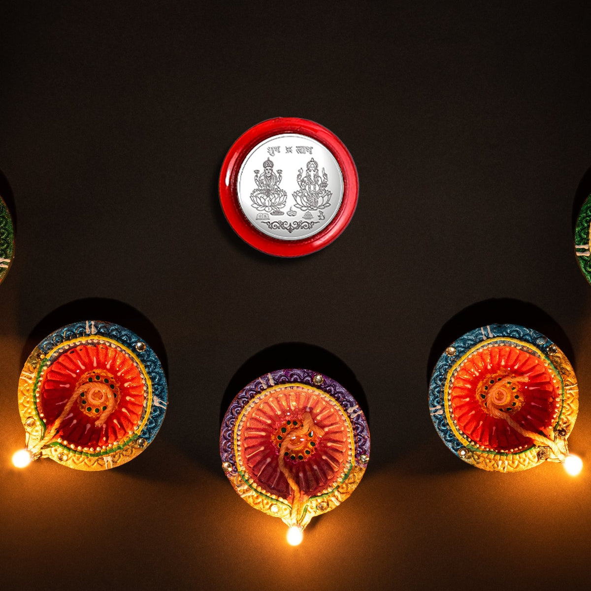 A silver coin with Lakshmi and Ganesha in a red case, surrounded by five lit colorful diyas on a dark background.