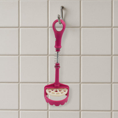 A pink and white plastic potato masher hangs vertically from a silver hook against a white tiled wall.