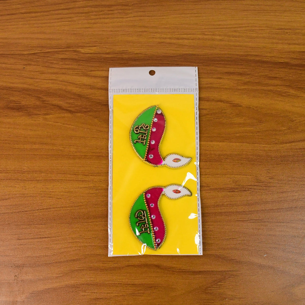Two diya-shaped stickers, pink and green with gold text, packaged in a clear plastic bag on a wooden surface.