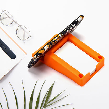 An orange plastic holder on a white desk, holding a smartphone vertically, with eyeglasses and a plant nearby.