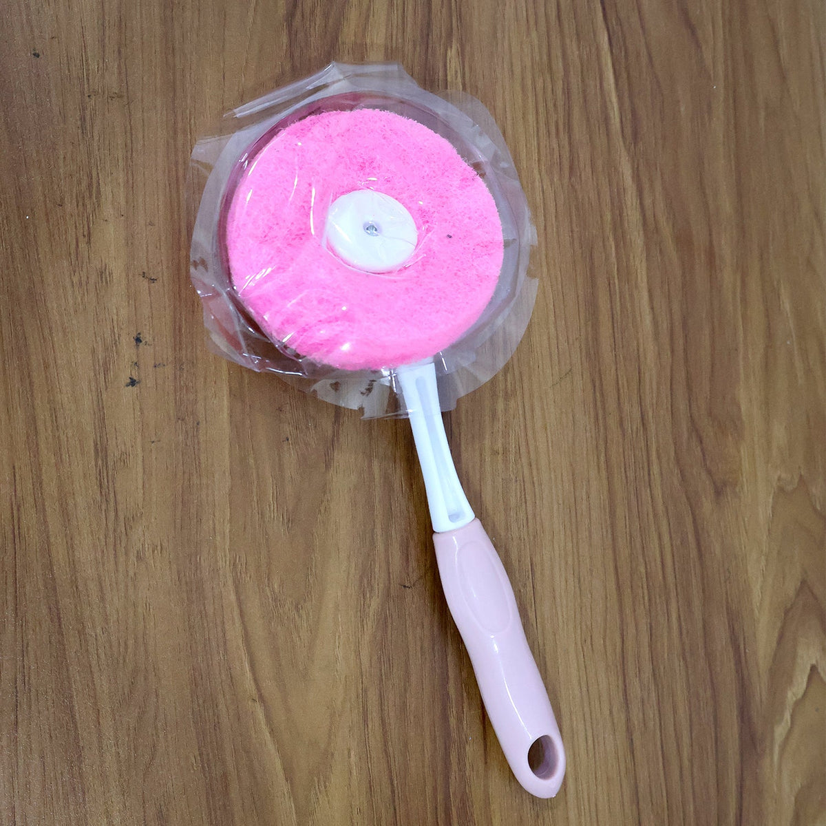 A pink and white long-handled dish scrubber, still in its clear plastic packaging, lies on a wooden surface.