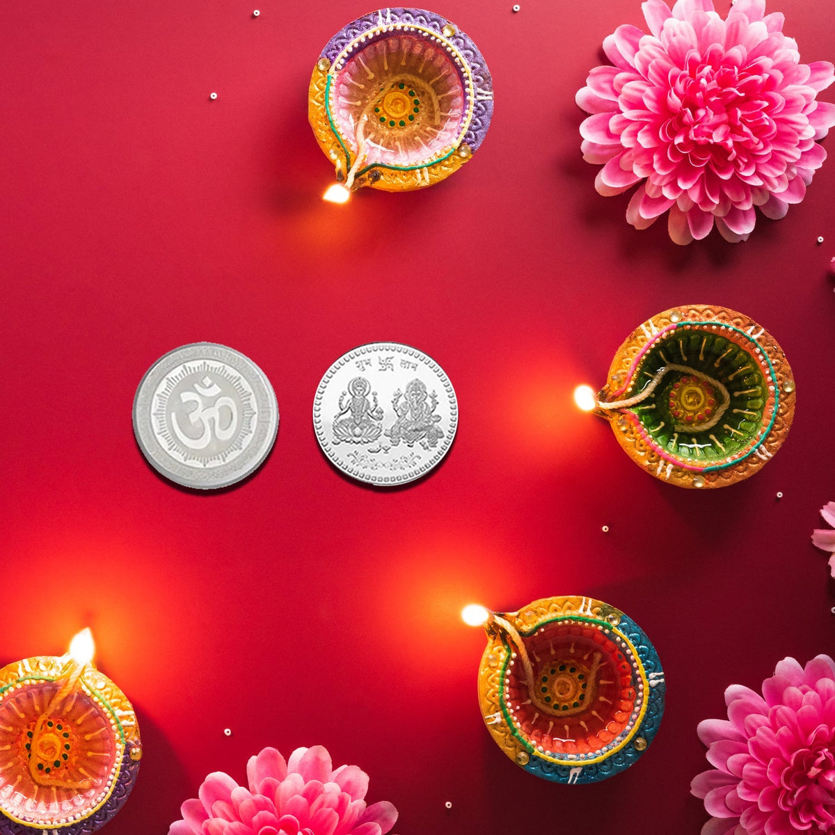 Two silver coins, one with Om and one with Lakshmi and Ganesha, surrounded by lit diyas and pink flowers on red.