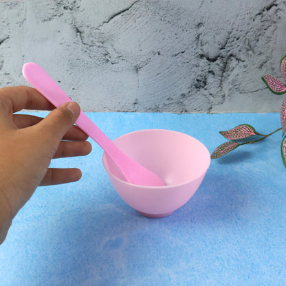 A hand holds a pink spatula in a pink mixing bowl, with a small pink measuring spoon on a blue surface.