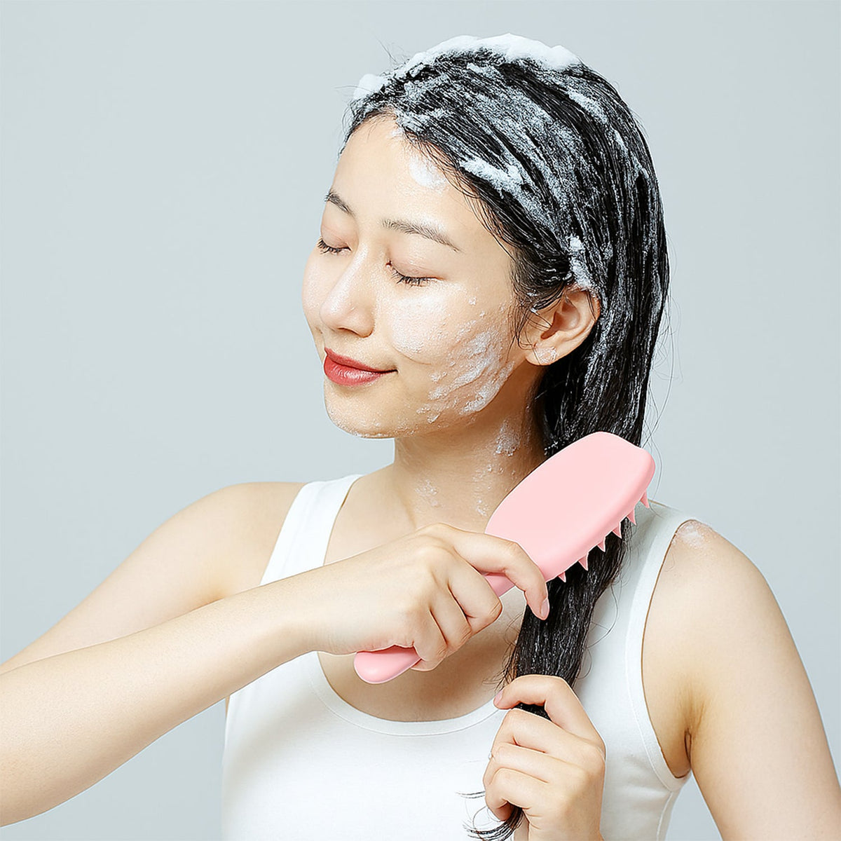 A woman with shampoo in her hair uses a pink silicone scalp massager brush.