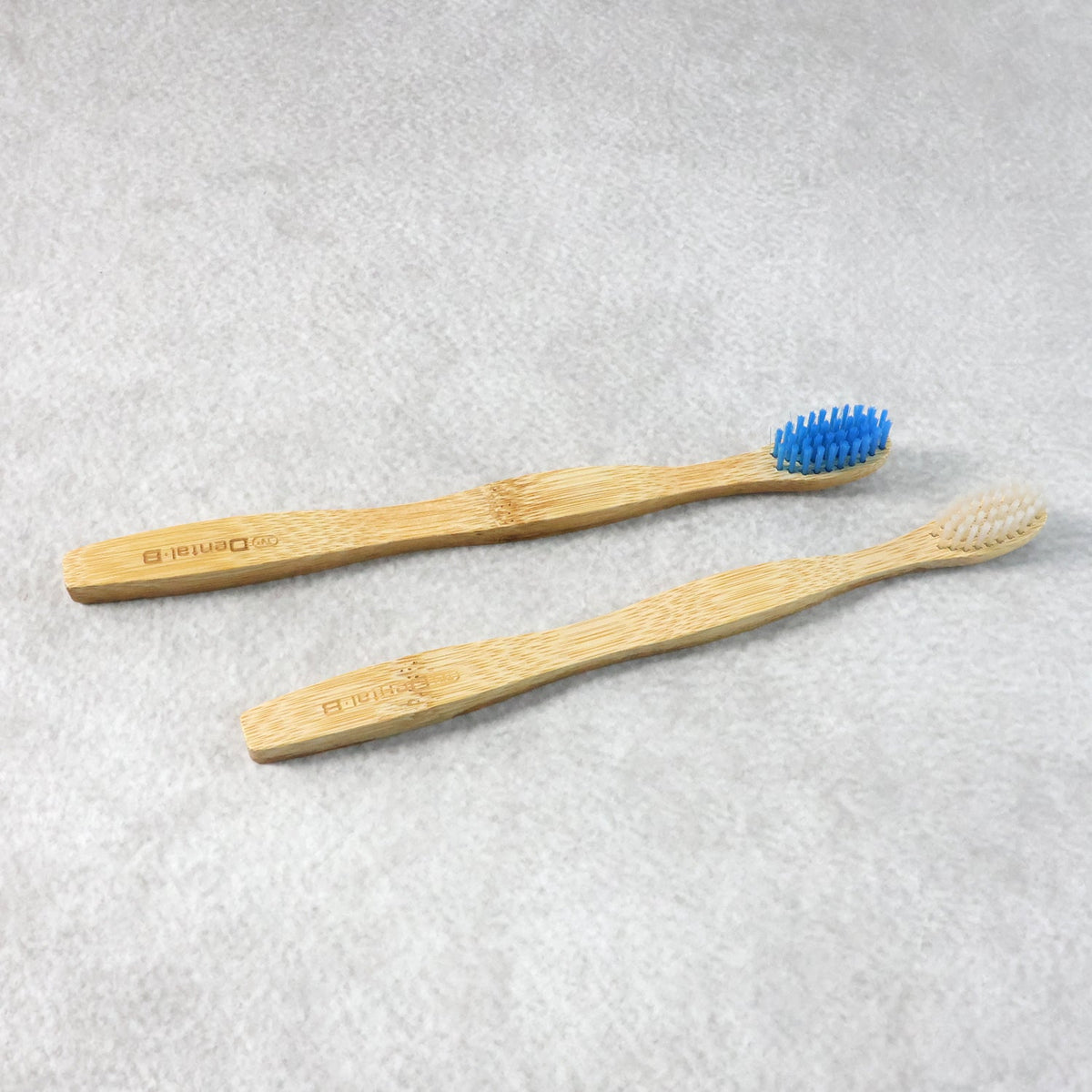 Two bamboo toothbrushes, one with blue bristles and one with beige bristles, lying on a light grey textured surface.