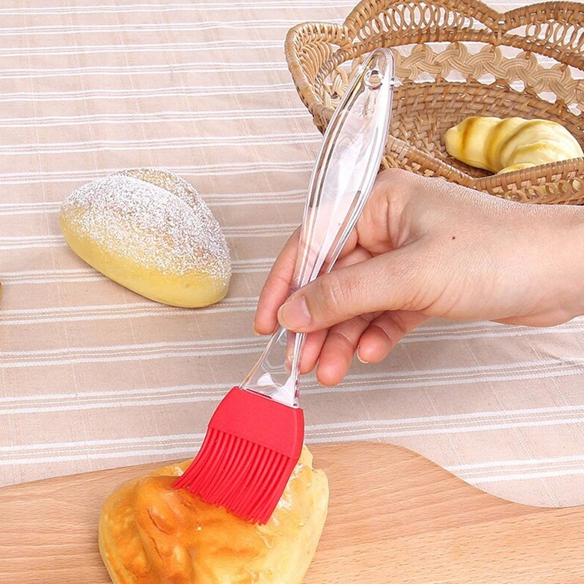 A hand uses a red silicone brush to glaze a golden-brown bun on a wooden cutting board next to a wicker basket.