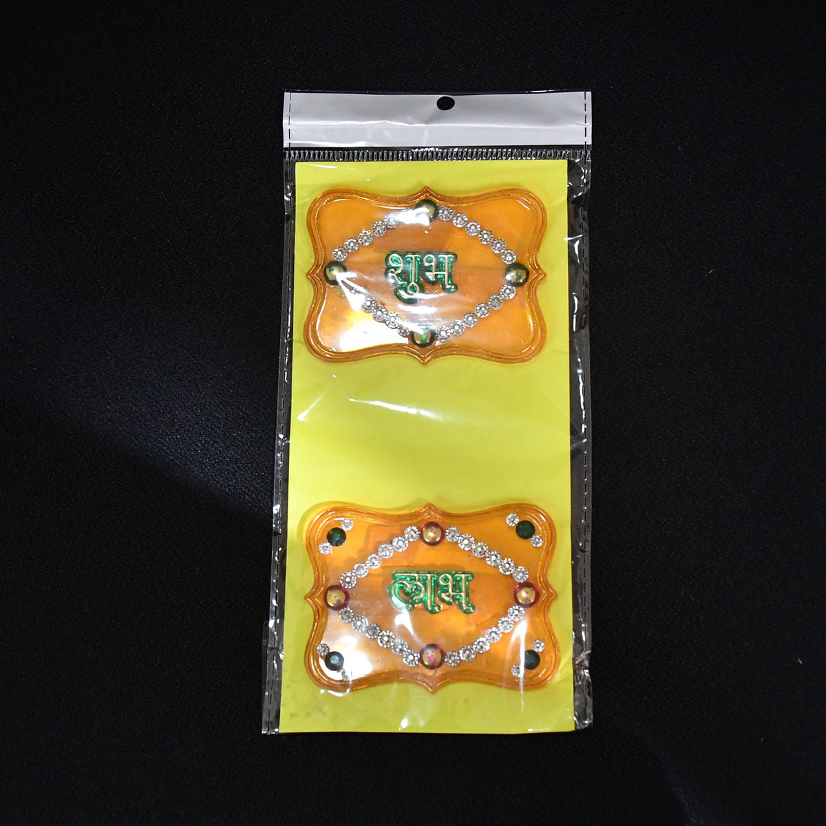Two yellow Shubh Labh wall hangings with green text and jewels, sealed in clear plastic packaging on a black background.