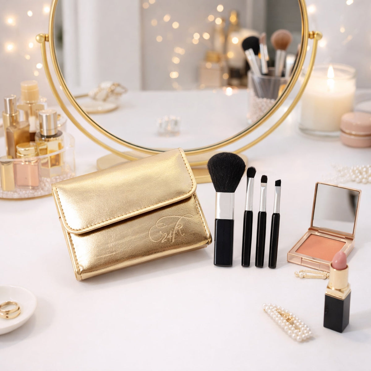 A gold makeup brush pouch and four brushes on a white vanity table with a round mirror and various cosmetics.