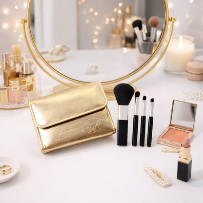 A gold makeup brush pouch and four brushes on a white vanity table with a round mirror and various cosmetics.