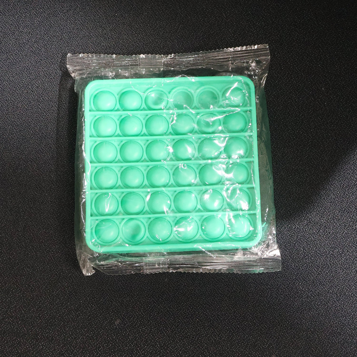 A square green silicone pop-it fidget toy sealed in clear plastic packaging on a dark surface.