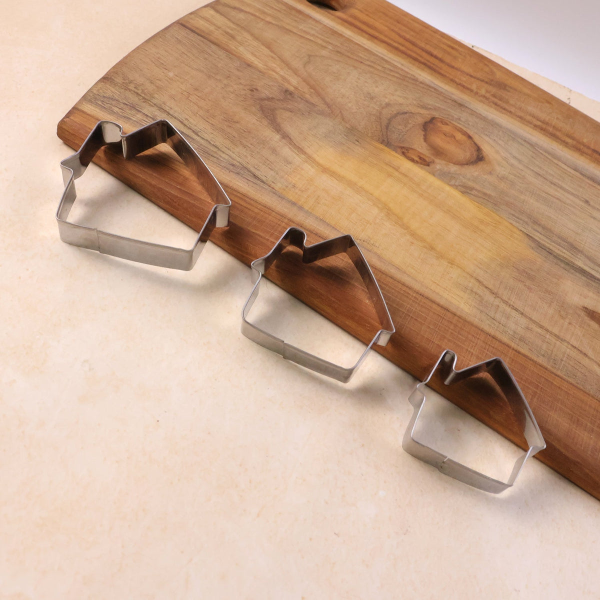 Three house-shaped stainless steel cookie cutters resting on a light-colored surface beside a wooden cutting board.