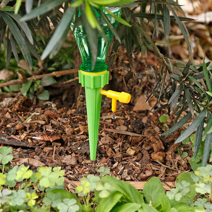 A green watering spike with a clear bottle inserted into a garden bed, with water dripping from the yellow valve.