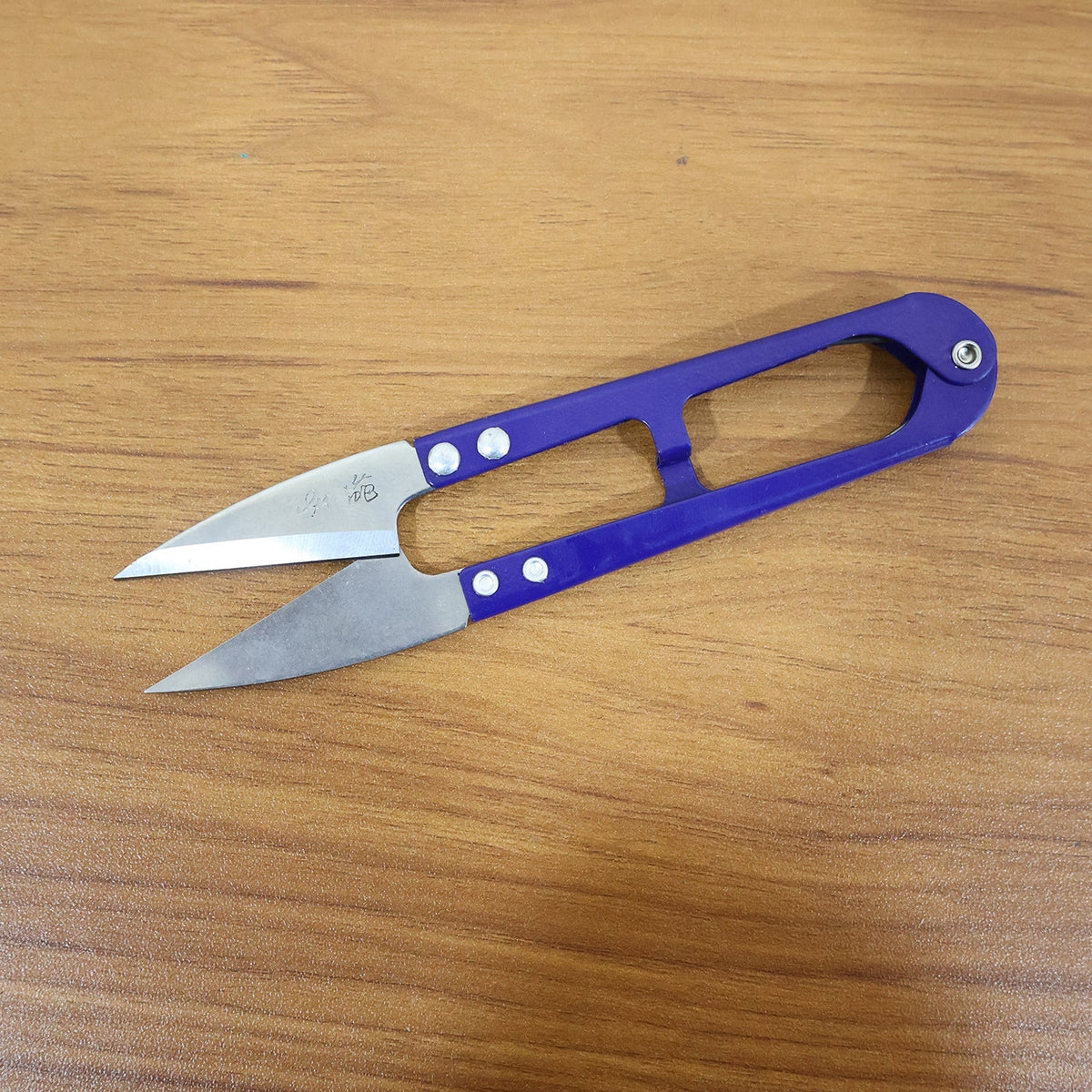 A pair of blue thread snips with silver blades, featuring some etched text, rests on a polished wooden surface.