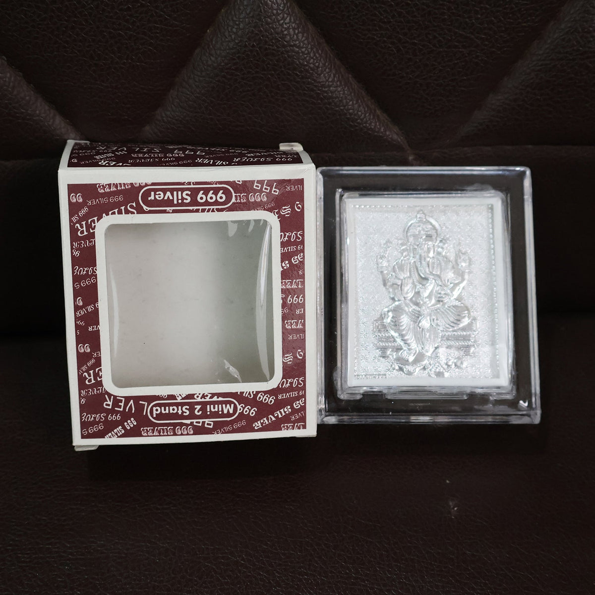 Silver-plated Ganesha idol in an acrylic frame next to its open retail box, both resting on a dark brown textured surface.