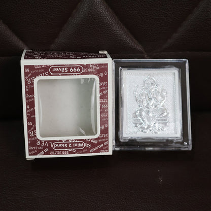 Silver-plated Ganesha idol in an acrylic frame next to its open retail box, both resting on a dark brown textured surface.