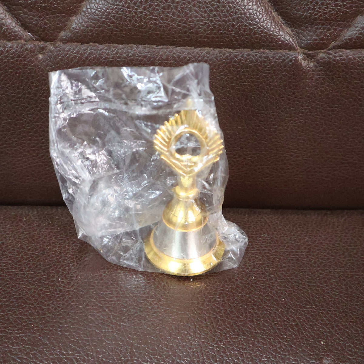 A small brass and steel pooja bell with a peacock feather design is sealed in a clear plastic bag on a brown textured