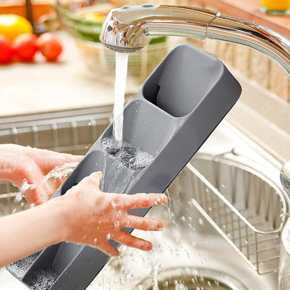 Two hands wash a gray plastic cutlery organizer under a chrome kitchen faucet, with water splashing into a sink.