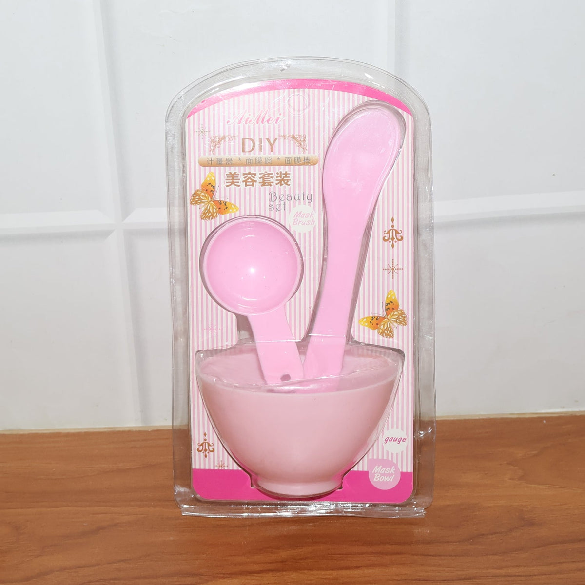 The pink facial mask mixing kit is presented in its clear plastic packaging, standing upright on a wooden surface.