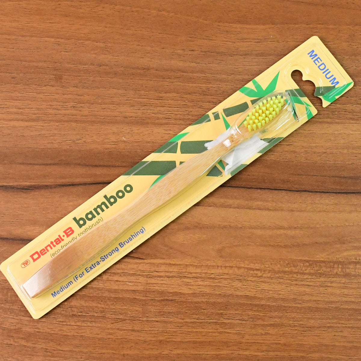 A packaged bamboo toothbrush with yellow bristles is displayed on a light brown wooden surface.