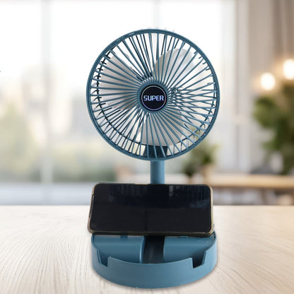 A teal portable fan with a phone resting in its base is on a wooden table, with a bright window in the background.