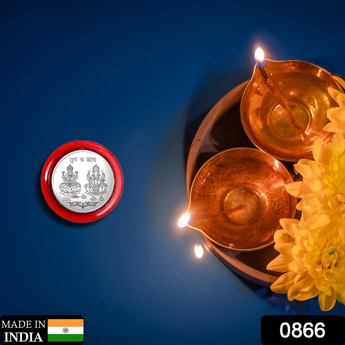 A silver coin with Lakshmi and Ganesha in a red case, next to two lit brass diyas and yellow flowers on blue.