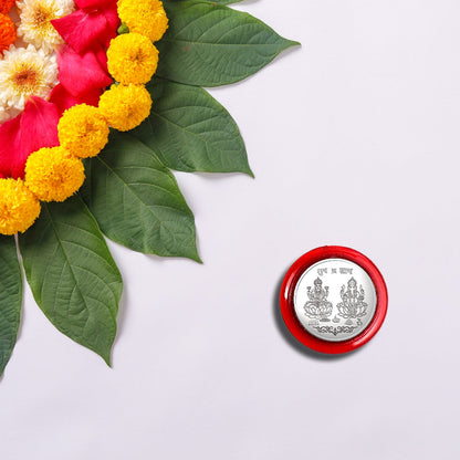 A silver coin with Lakshmi and Ganesha in a red case, beside a floral rangoli design with green leaves and yellow flowers.