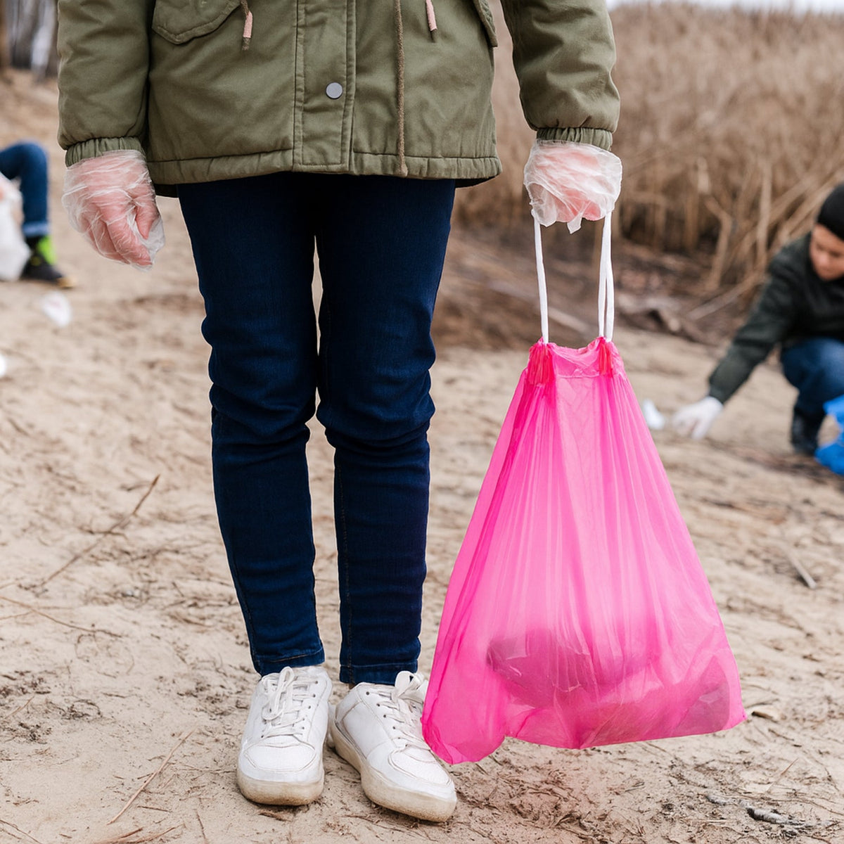 A person wearing gloves holds a full pink drawstring garbage bag on a sandy beach