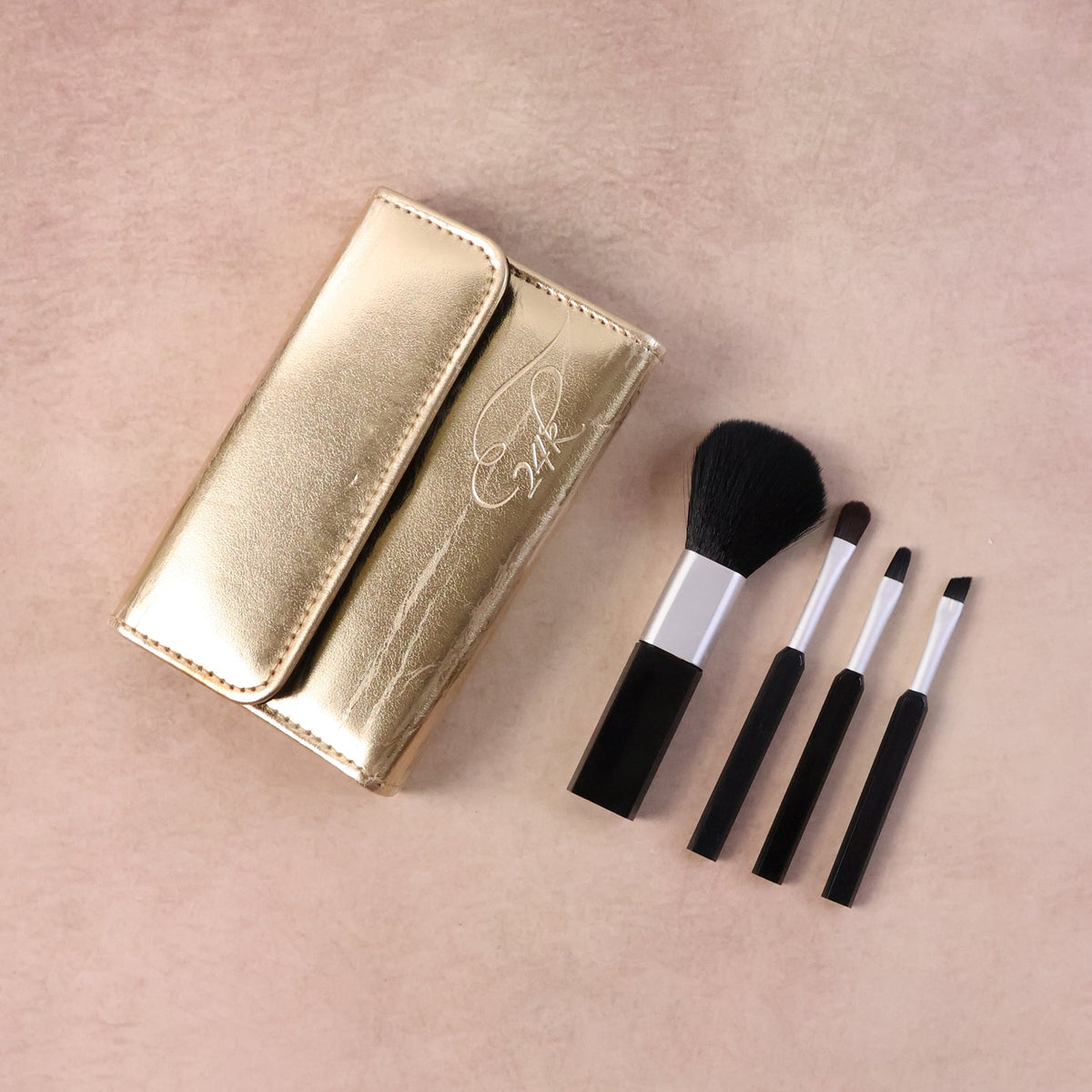 A gold makeup brush pouch with '24K' embossed and four black and silver brushes on a light pink surface.