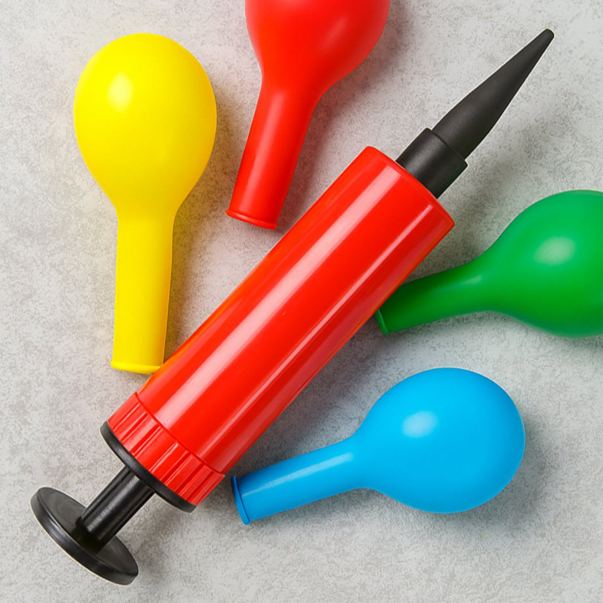 A red and black manual balloon pump surrounded by uninflated yellow, red, green, and blue balloons on a gray surface.