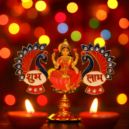 A Lakshmi idol sits between the two peacock Shubh Labh decorations, with lit oil lamps and colorful bokeh lights.