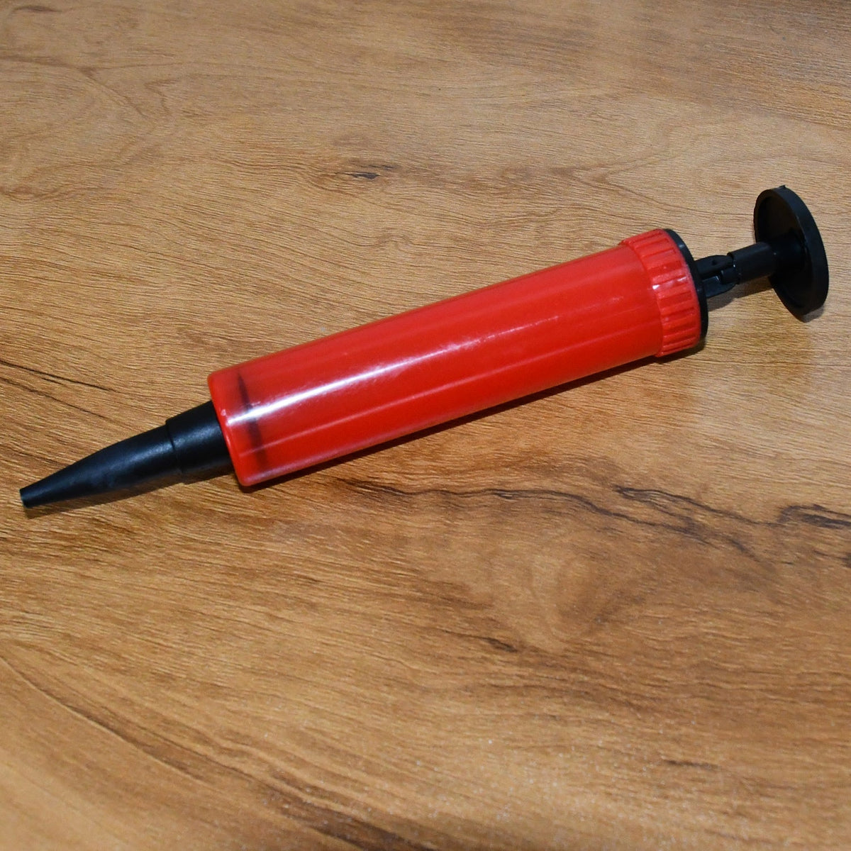 A red and black manual balloon pump rests horizontally on a warm-toned wooden surface.