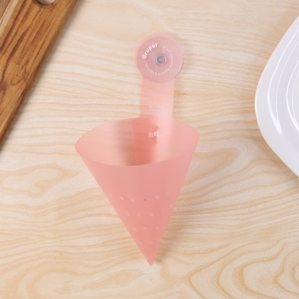 A pink silicone sink strainer with a suction cup, empty, on a light wood countertop next to a white plate.