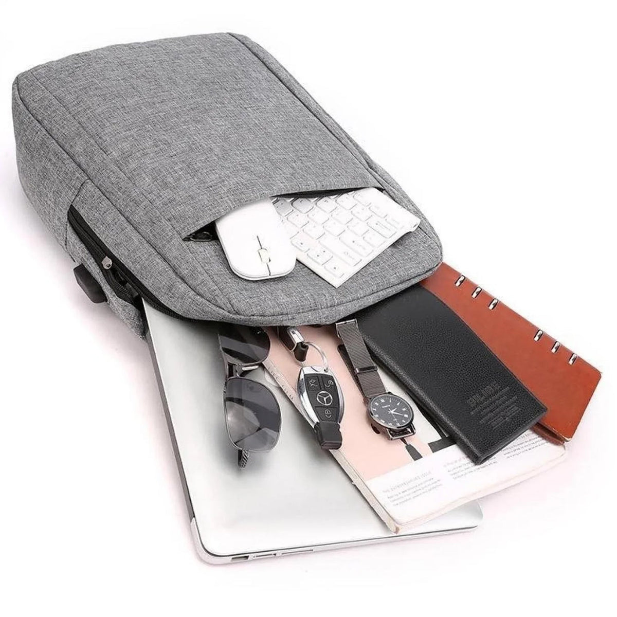 A grey backpack is open, revealing a laptop, mouse, keyboard, sunglasses, watch, and notebooks spilling out.