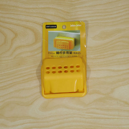 A yellow plastic sponge holder in its original packaging, lying on a light wooden surface.