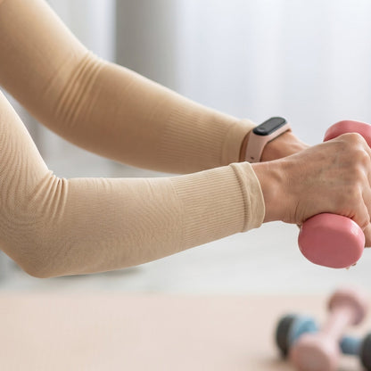 Close-up of a woman's arms wearing beige arm sleeves and a smartwatch, holding pink dumbbells.