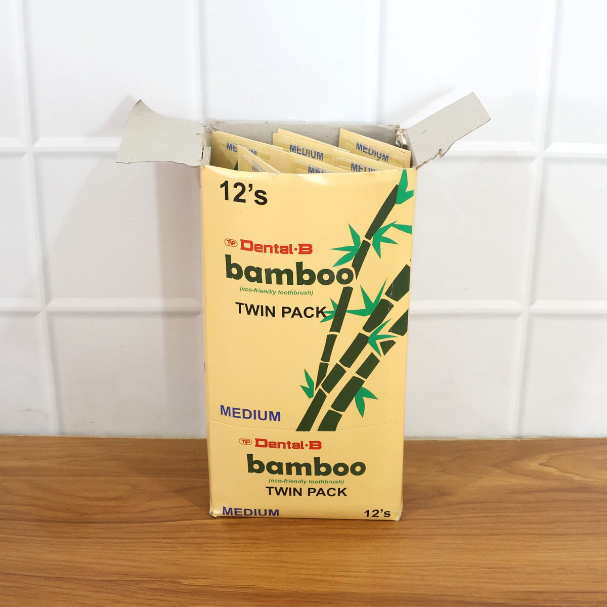 An open cardboard box filled with multiple twin packs of bamboo toothbrushes, against a white tiled wall.