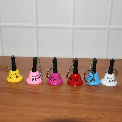 Six mini hand bell keychains in different colors, each with a unique phrase, lined up on a dark wooden surface.