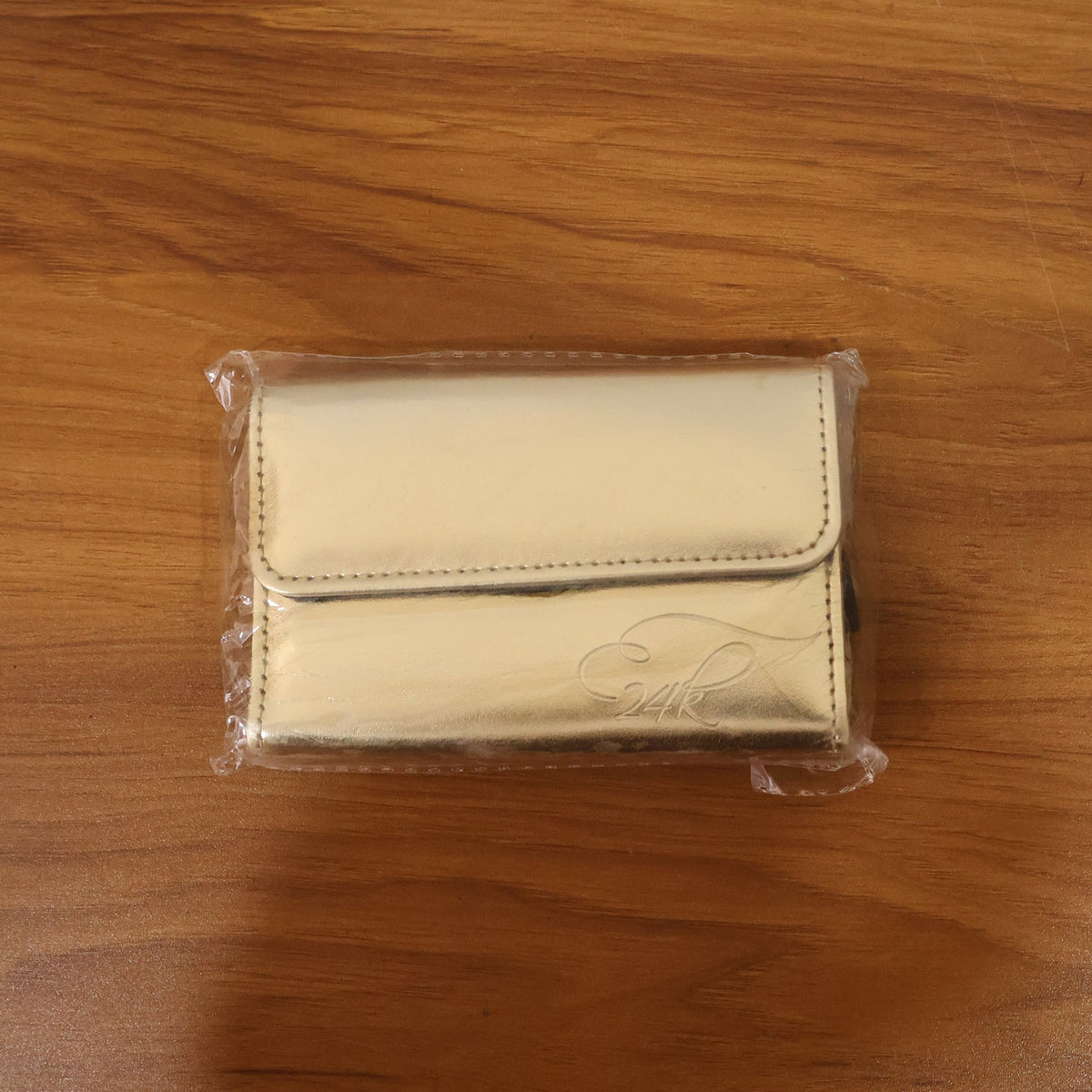 A gold makeup brush pouch, sealed in clear plastic, resting on a light brown wooden surface.