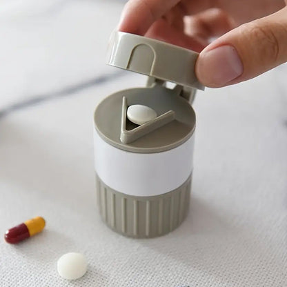 A hand opening a gray and white pill cutter with a white pill inside and a capsule nearby.