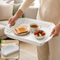 A person holds a white foldable serving tray with toast, tea, and fruit on it, in a living room.